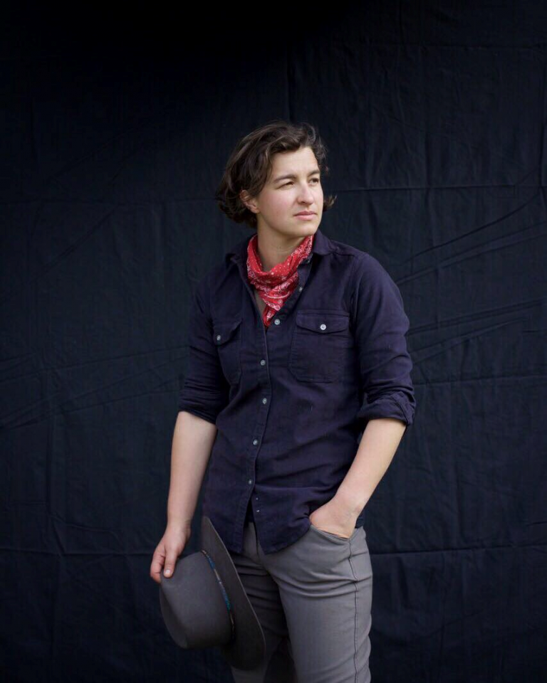 Person wearing a navy button-up shirt and red bandana stands against a dark backdrop, holding a gray hat in one hand.