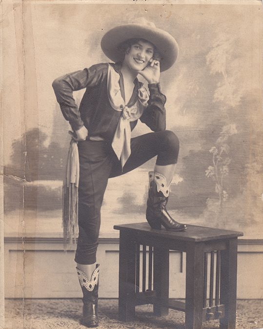 Vintage photo of a smiling woman in a Western costume, wide-brimmed hat, scarf, fringe sash, and tall boots with decorative tops, one foot resting on a small table.