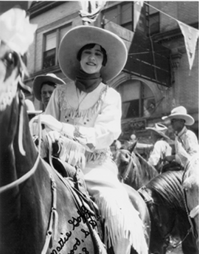 Woman in fringed Western dress and wide-brim hat rides a horse in a street parade; other riders and bunting visible in the background.