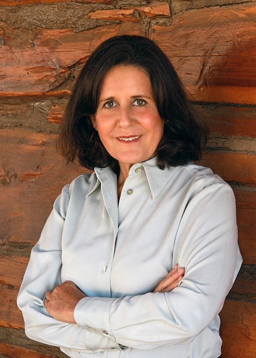 Professional woman with dark hair, wearing a light blue button-down shirt, standing against a wooden wall with arms crossed and a slight smile.