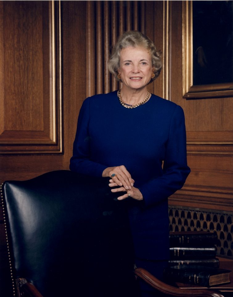 Portrait of a smiling older woman in a royal blue dress, standing in a wood-paneled room with books nearby.