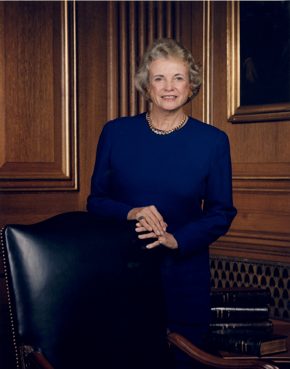 Portrait of a smiling older woman in a royal blue dress, standing in a wood-paneled room with books nearby.