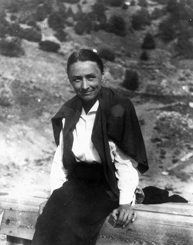 Black-and-white photo of a woman sitting on a wooden railing outdoors, wearing a white blouse and dark skirt with a jacket draped over her shoulders, hillside behind her.