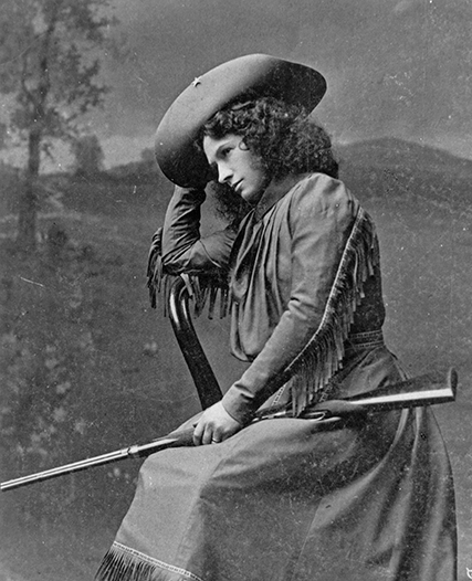 Seated woman in period dress with fringe, wearing a wide-brim hat, resting a rifle on her lap.