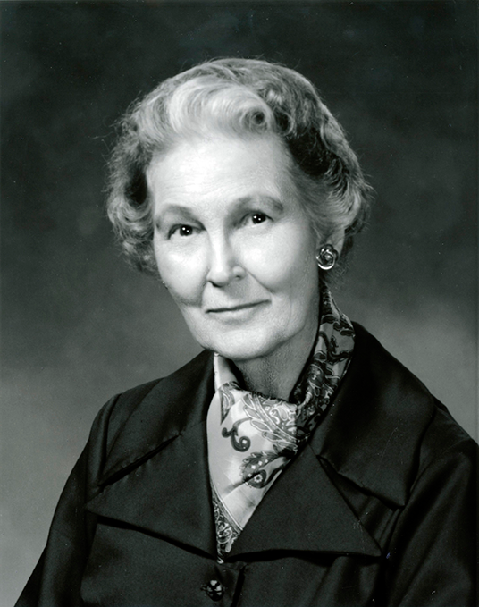 Portrait of an elderly woman with short curled hair, wearing a dark blazer and patterned scarf, looking at the camera with a gentle smile.