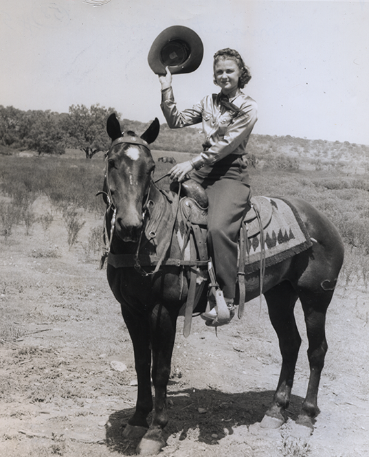 Woman riding a horse in a dusty rural landscape, tipping her hat toward the camera while smiling (vintage photo).
