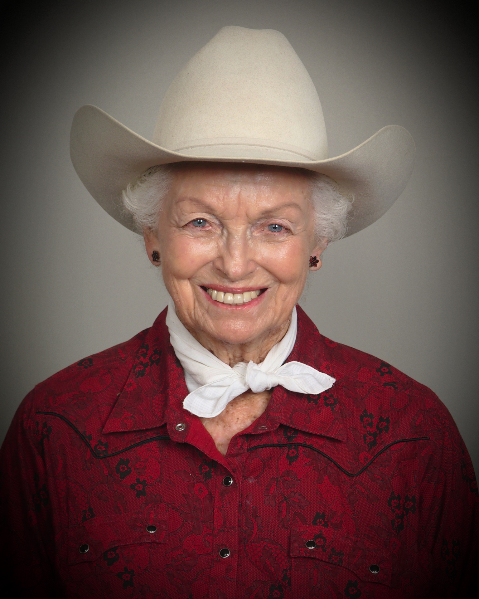Older woman wearing a white cowboy hat, red lace shirt, and white neck scarf, smiling warmly at the camera.