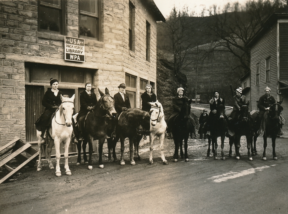 Line of riders on horses along a dirt street in front of a stone library with a WPA 'Pack Horse Library' sign and USA.