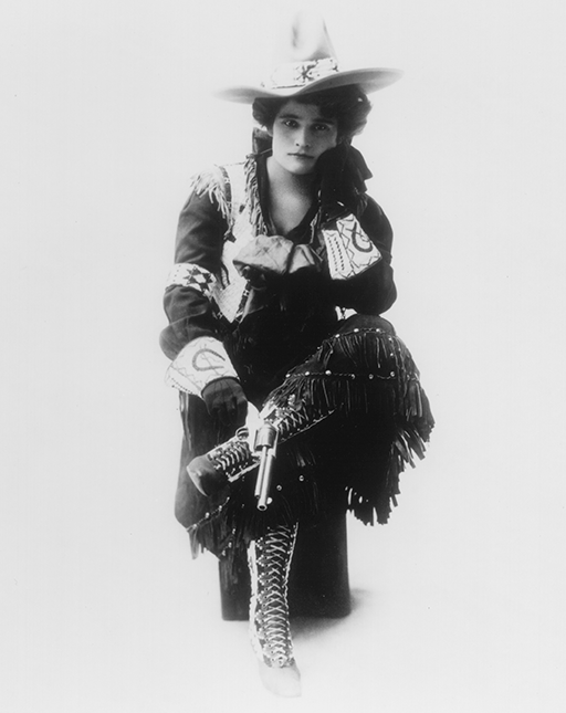 Woman in a fringed Western outfit with a wide hat, sitting and holding a pistol.