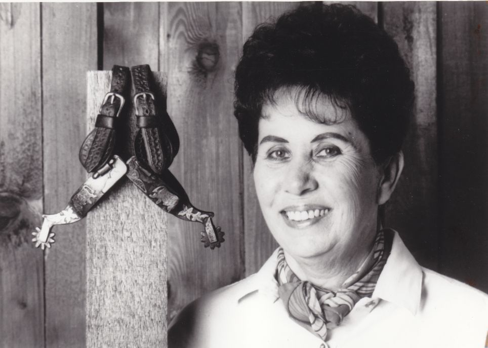 Black-and-white photo of a smiling woman in a light blouse with a scarf, standing beside a wooden post that has two decorative leather straps or harness pieces hanging from it.