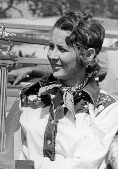 Smiling woman in western shirt and scarf stands beside a tall trophy outdoors, likely at an award ceremony.