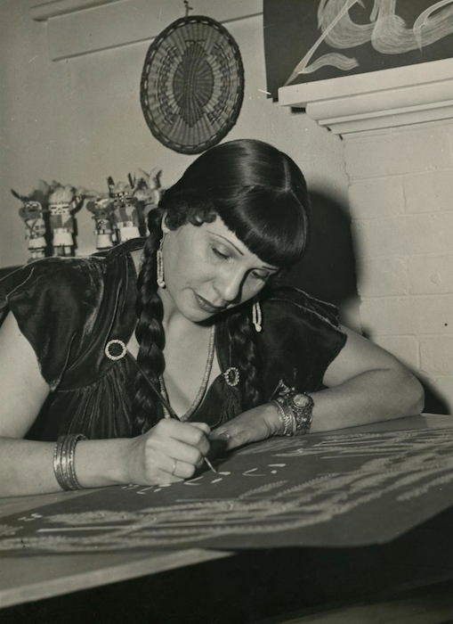 Black-and-white photo of a woman with braided hair, sitting at a table and writing on a large surface with chalk or marker, wearing jewelry and a dark blouse in a vintage setting.