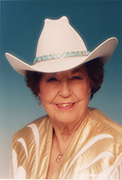 Smiling elderly woman in a white cowboy hat with turquoise band and gold blouse against a blue background.