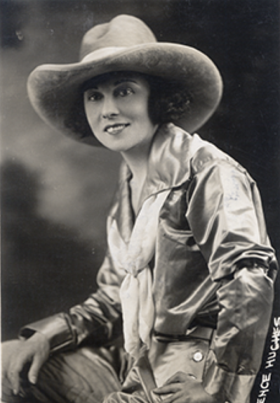 Black-and-white portrait of a smiling woman wearing a wide-brimmed hat and satin blouse, posed seated with one hand resting on her knee.