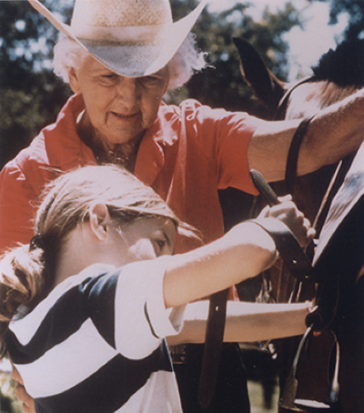 An elderly woman wearing a straw hat helps a young girl saddle a horse outdoors.