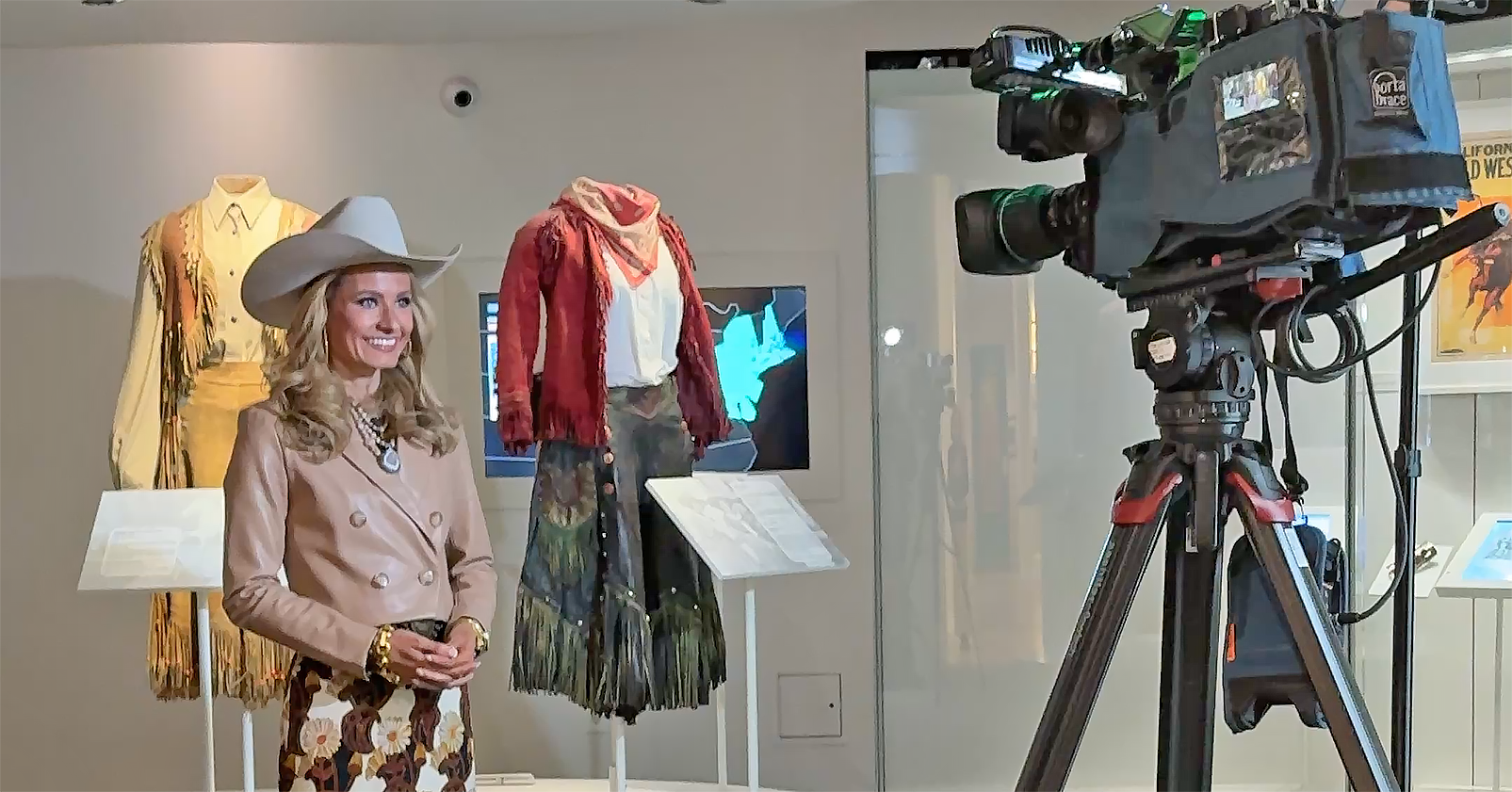 Life-size mannequin wearing a beige suit and wide-brim hat in a museum display, with vintage costumes and a filming camera on a tripod nearby.