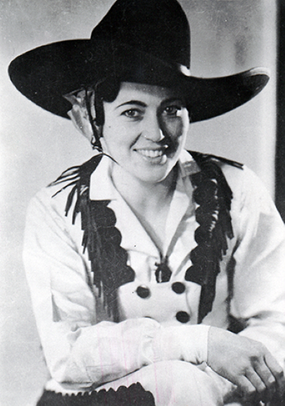 Smiling woman in a wide-brimmed cowboy hat and white shirt with fringe, posing for a portrait.