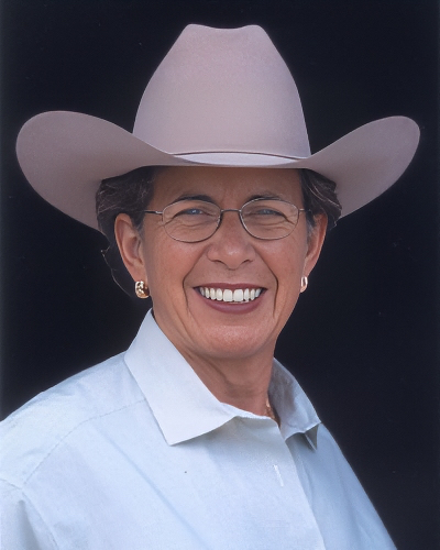 Smiling woman wearing a light-colored cowboy hat, glasses, gold earrings, and a light blue button-down shirt.
