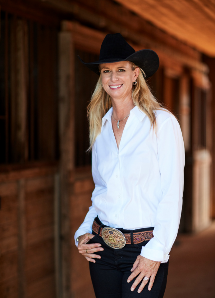 Smiling woman in a white button-up shirt, black jeans, and a large western belt buckle, wearing a black cowboy hat in a rustic wooden setting