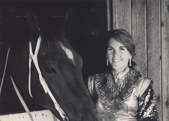 Smiling woman in a rhinestone western shirt standing beside a horse at a stable door.