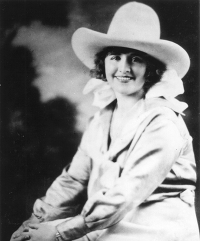 Black-and-white portrait of a smiling woman wearing a wide-brimmed hat and light dress, seated and facing the camera.