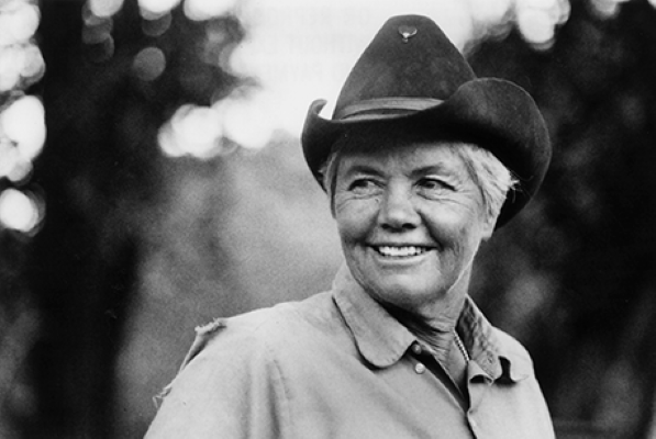 Smiling woman wearing a cowboy hat and light jacket outdoors, looking to the side.