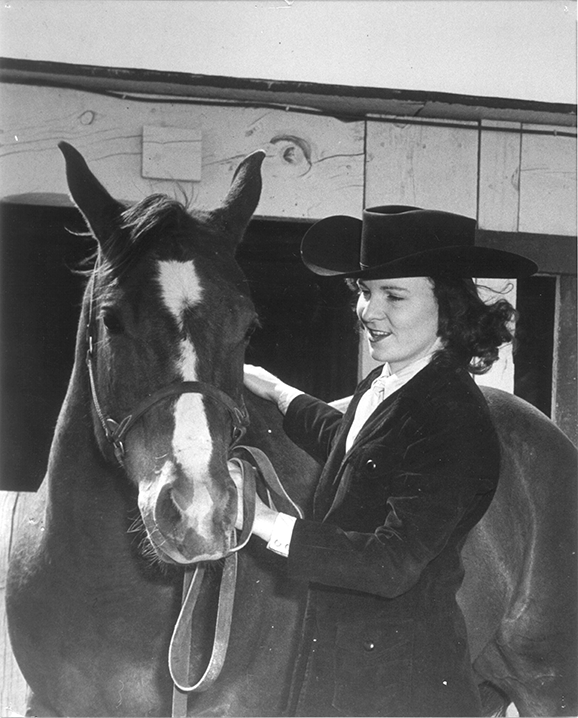 Woman in a dark suit and wide-brimmed hat standing beside a horse in a stable, gently holding its reins.