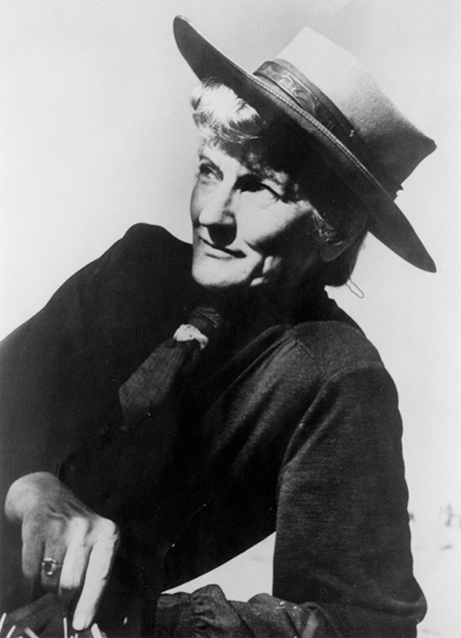 Black-and-white portrait of an older woman in a wide-brimmed hat and dark sweater, smiling slightly.