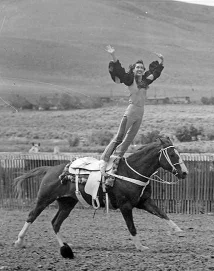 Woman performing a trick ride, standing on a galloping horse with arms raised in an outdoor arena, wearing western-style attire.