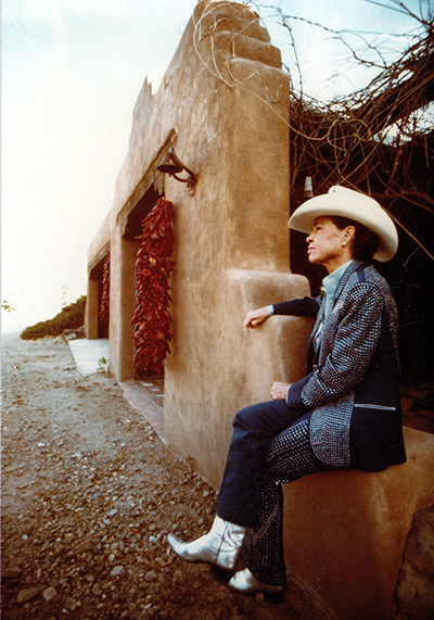 Older man in a white cowboy hat sits on an adobe wall beside a doorway framed by hanging red chili peppers, in a desert setting.