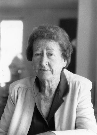 Portrait of an elderly woman with short, wavy hair, wearing a light blazer over a dark blouse, indoors.