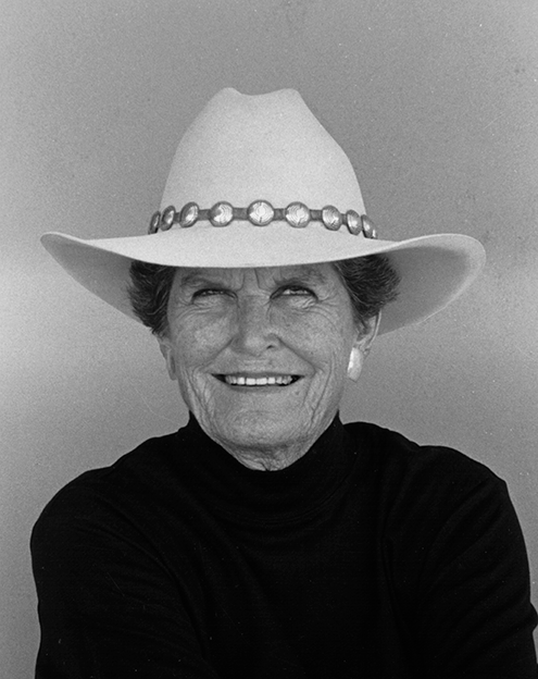 Smiling elderly woman in a wide-brimmed hat with a beaded band, wearing a black turtleneck.