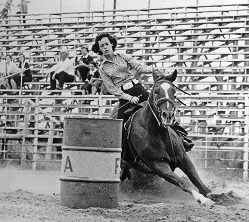 Female rider leans into a turn on a galloping horse as it rounds a barrel in a rodeo arena; stands of spectators in the background.