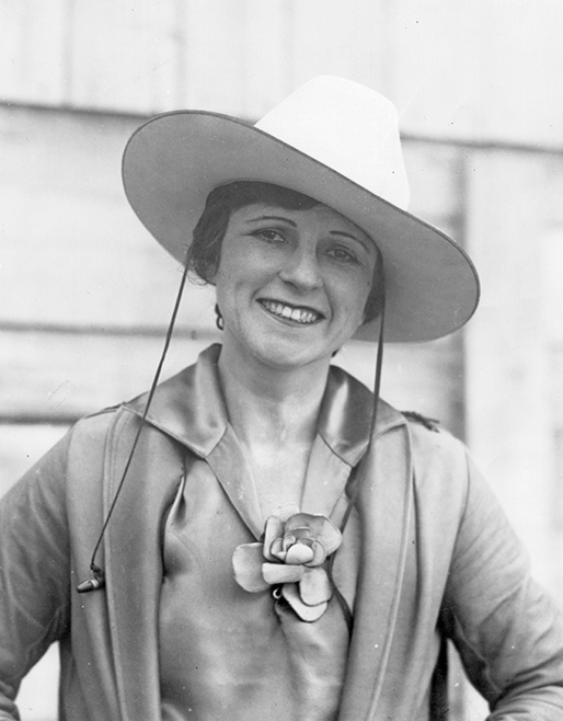 Black-and-white photo of a smiling woman wearing a wide-brimmed hat and a satin blouse with a floral brooch.