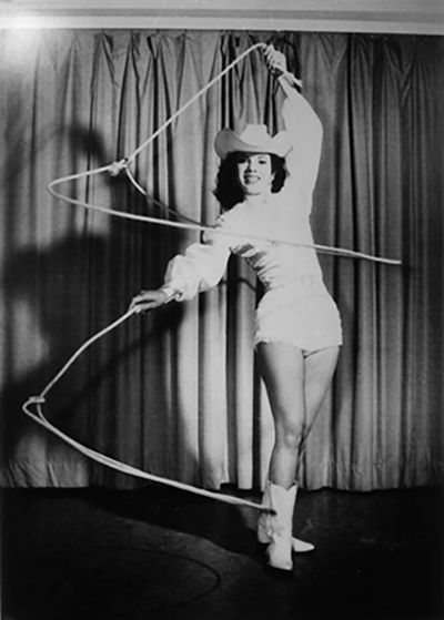 Black-and-white photo of a woman in a cowboy outfit spinning a lasso on a stage with a curtained backdrop.