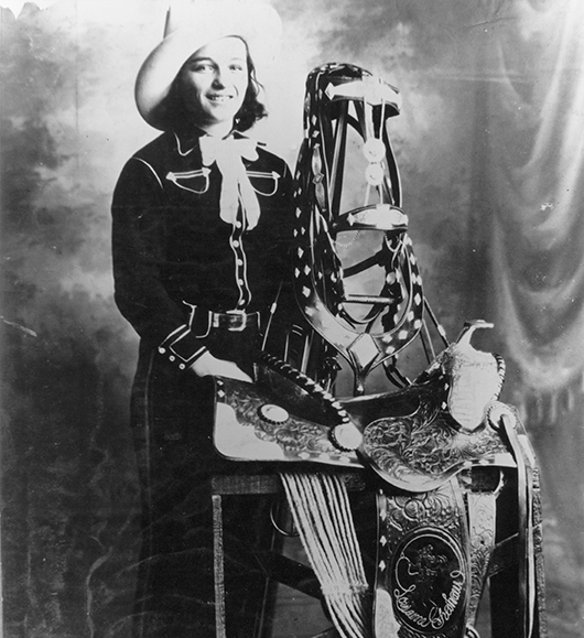 Smiling woman in a western outfit stands beside an ornately decorated horse saddle and tack on a chair in a vintage studio photo.