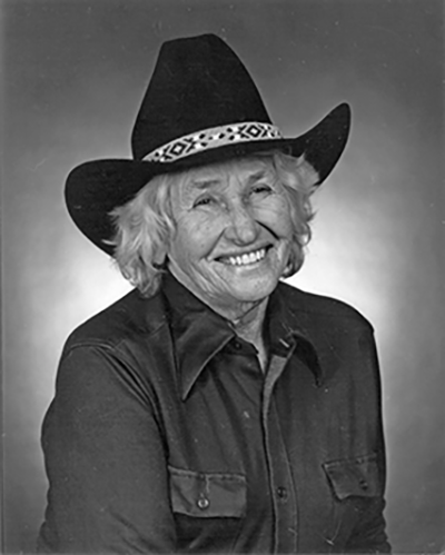 Smiling older woman wearing a dark western-style shirt and a cowboy hat with a patterned band.