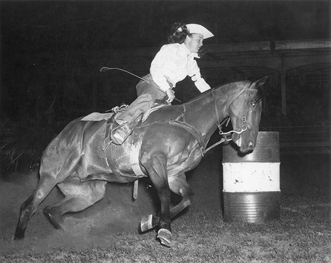 A rider in a light shirt and cap leans forward on a horse as it jumps over a barrel obstacle at a nighttime equestrian event.