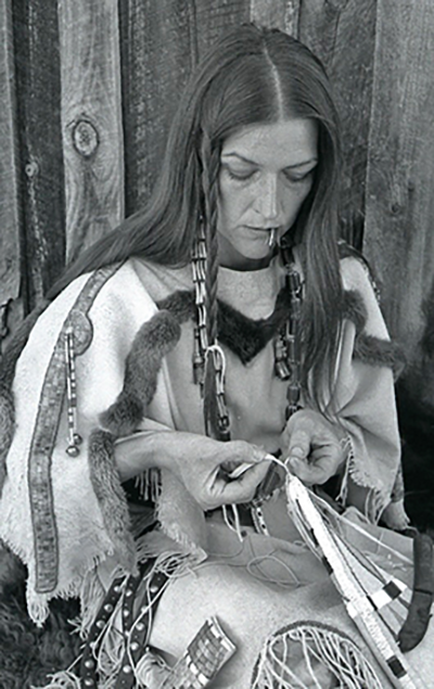 Native American woman in fringed traditional clothing sits against a wooden fence, sewing leather strips with a cigarette in her mouth.
