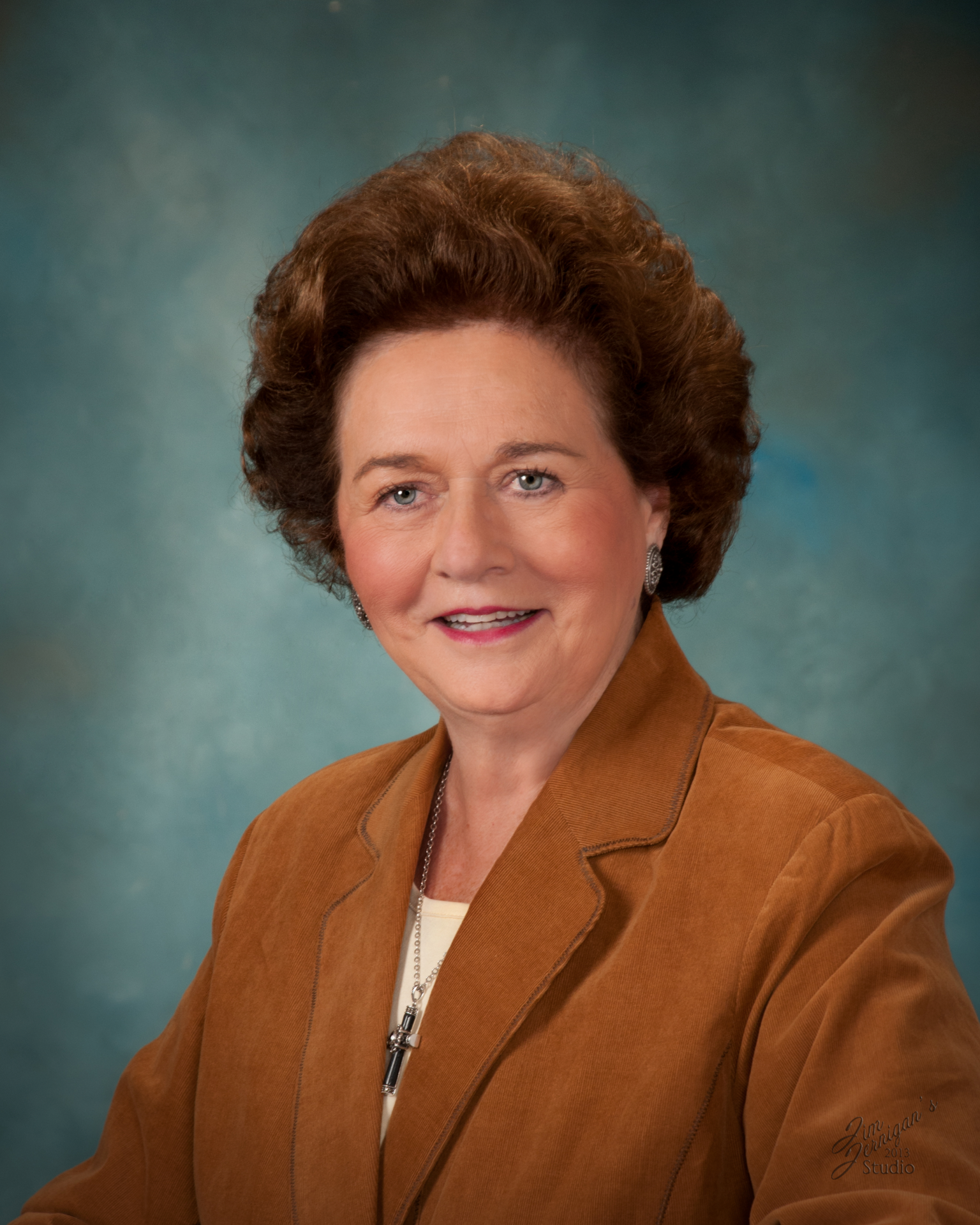Head-and-shoulders portrait of a smiling older woman with short brown hair, wearing a brown suede jacket and a necklace against a teal studio backdrop.