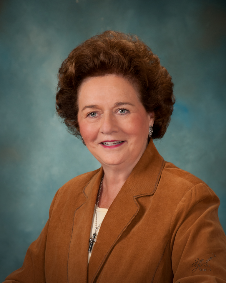 Head-and-shoulders portrait of a smiling older woman with short brown hair, wearing a brown suede jacket and a necklace against a teal studio backdrop.