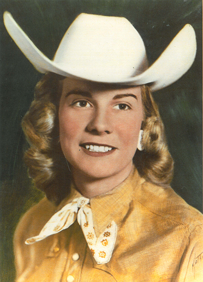 Portrait of a smiling woman in a white cowboy hat and yellow western shirt, tied scarf, posed against a dark backdrop.