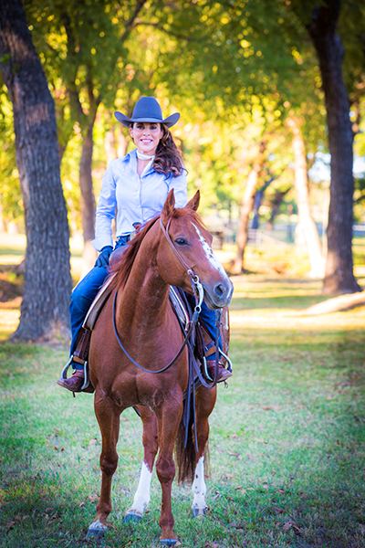 Smiling woman in a blue shirt and cowboy hat riding a brown horse through a sunlit park with trees.