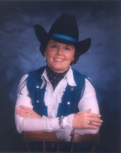 Smiling woman in a black cowboy hat with a blue band, blue vest, white ruffled blouse, seated with arms crossed on a chair back.