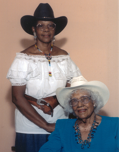 Two women with cowboy hats posing indoors: a standing woman in a white off-the-shoulder blouse and belt, and an seated older woman in a blue blazer and white hat.