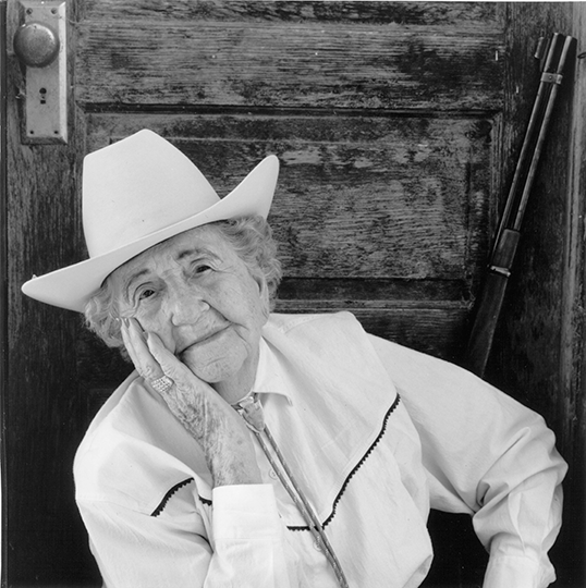 Elderly woman in a white cowboy hat rests her head on her hand, smiling softly beside a weathered wooden door.