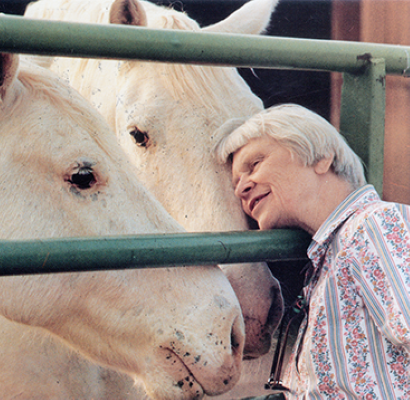 Older man gently leans in to nuzzle two white horses through a green fence at a stable.