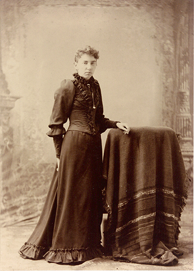 Sepia-toned portrait of a Victorian-era woman in a high-collared, ruffled dress standing beside a draped table.