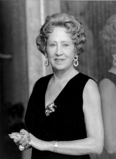 Older woman in a sleeveless black evening gown with a jeweled brooch at the chest, large drop earrings, and a bracelet, standing with hands clasped.