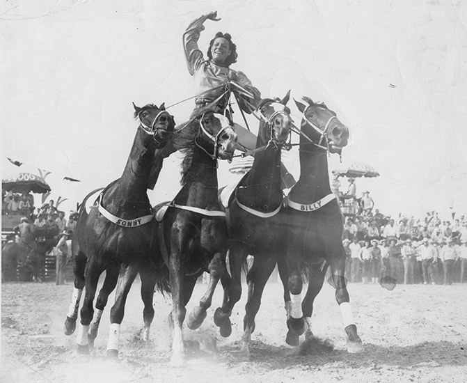 Rider atop a four-horse sulky team leading a race, with cheering crowd in the background.